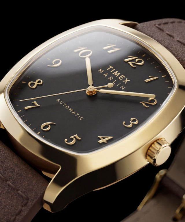 Närbild av guldfärgade Timex Marlin Chatham Automatic som visar dess svarta urtavla, applicerade guldfärgade arabiska siffror, guldfärgade visare och bruna läderarmband