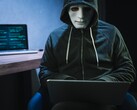 En maskerad person med huva arbetar på en bärbar dator i ett mörkt rum.