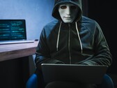 En maskerad person med huva arbetar på en bärbar dator i ett mörkt rum.