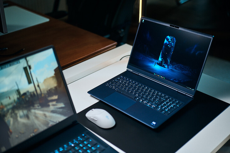 En mörkblå gaming-laptop i mellanklassen (Alienware Aurora 16X) vinklad på en skrivbordsunderlägg med en lysande blå sci-fi-bakgrundsbild, placerad bredvid en vit trådlös mus med en annan laptop suddig i förgrunden.