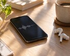 Smartphone som visar en ljudvågform bredvid trådlösa öronsnäckor och en kaffekopp på ett skrivbord av trä