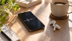 Smartphone som visar en ljudvågform bredvid trådlösa öronsnäckor och en kaffekopp på ett skrivbord av trä