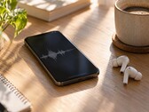 Smartphone som visar en ljudvågform bredvid trådlösa öronsnäckor och en kaffekopp på ett skrivbord av trä