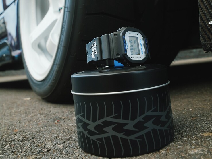 En Casio G-Shock DW5600TT25-1 x Toyo Tires-klocka utställd på en metallburk med däckmotiv bredvid ett bilhjul.