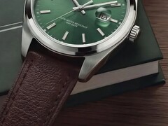 Närbild av klockan Timex Legacy Day and Date som visar dess gröna urtavla med solstrålemönster, silverfärgade boettring och borstade bandhorn, räfflade krona och strukturerade bruna läderarmband mot en mörkgrön ask och en träyta.