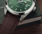 Närbild av klockan Timex Legacy Day and Date som visar dess gröna urtavla med solstrålemönster, silverfärgade boettring och borstade bandhorn, räfflade krona och strukturerade bruna läderarmband mot en mörkgrön ask och en träyta.