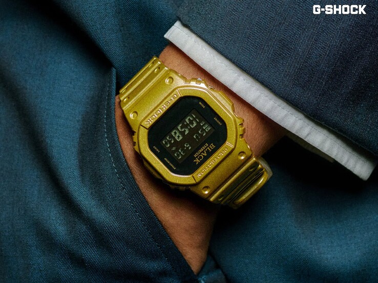 Närbild av en guldfärgad Casio G-Shock DW-5600BEP-9-klocka som bärs på en handled mot mörkblått tyg. Klockan har en svart digital display, guldfärgat boett och armband samt BlackEyePatch-logotypen på bezeln och urtavlan.