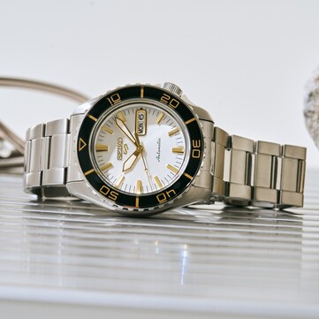 Seiko 5 Sports SKX HDB004-klocka med silvervit urtavla, svart vridbar bezel med guldfärgade markeringar, guldfärgade visare och index samt armband i rostfritt stål, fotograferad i vinkel.