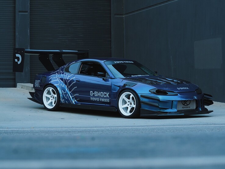 Blå driftbil med grafik från G-Shock och Toyo Tires parkerad utanför en industribyggnad.