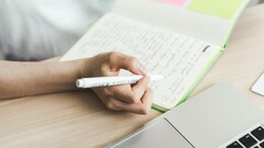 En person skriver anteckningar i en anteckningsbok bredvid en öppen bärbar dator