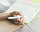 En person skriver anteckningar i en anteckningsbok bredvid en öppen bärbar dator