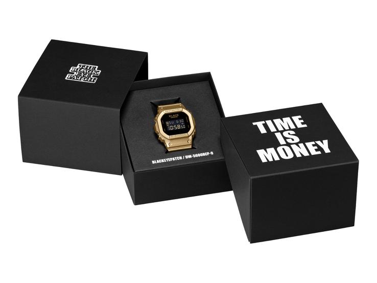 En guldfärgad Casio G-Shock DW-5600BEP-9-klocka med svart digital display ligger mellan två svarta presentförpackningar. Klockan har BlackEyePatch-logotypen på urtavlan, medan det på förpackningen i förgrunden står ”TIME IS MONEY”.