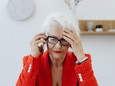 En äldre kvinna med vitt hår och glasögon som pratar i telefon i ett ljust vardagsrum, med handen mot tinningen.