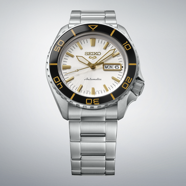 Seiko 5 Sports SKX HDB004 med silvervit urtavla, svart vridring med guldfärgade markeringar, guldfärgade visare och index samt länk i rostfritt stål, visad mot en ljusgrå bakgrund.